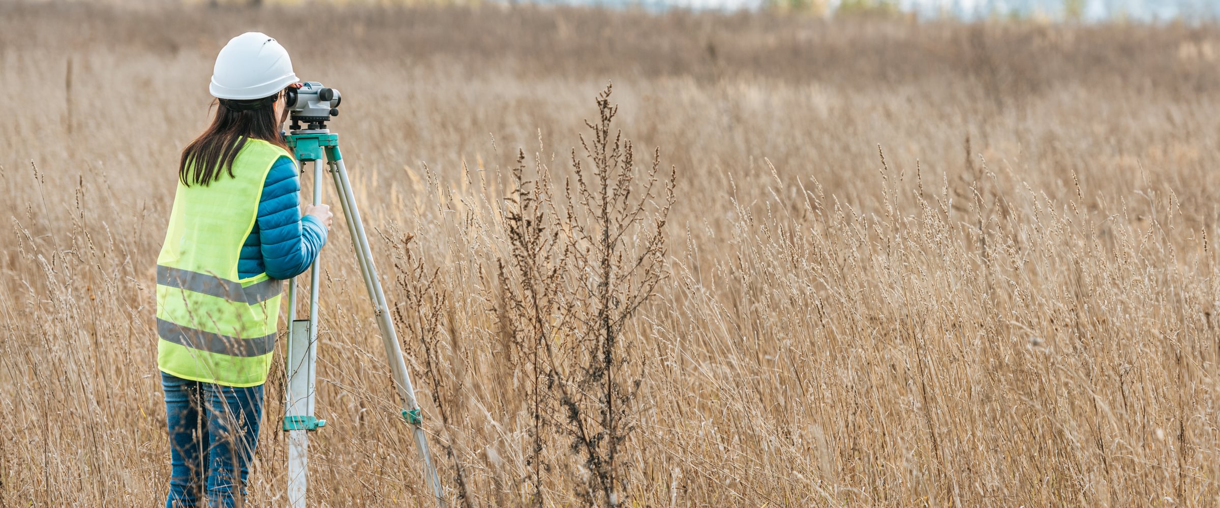 Surveyor in a hard hat and safety vest using a tripod-mounted level in a field of tall dry grass, with a tree line in the distance.
